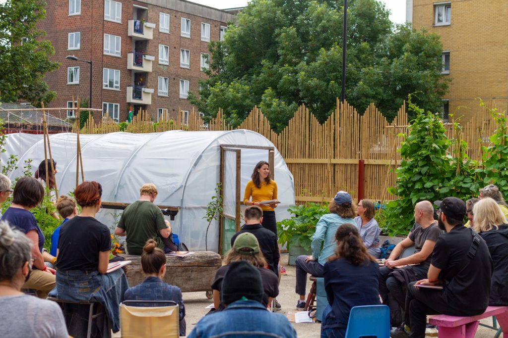 A group of around 20 people are sitting in a circle in a garden, facing a woman who is standing in front of a polytunnel, explaining the workshop plan.