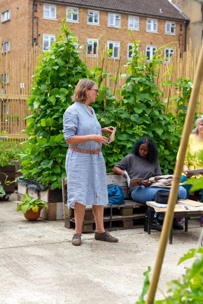 A woman stands with a booklet in her hand, in an urban garden, with a residential building behind her, and a concrete floor. Behinds her are bean plants growing up from raised beds. Another woman sits behind her on some pallet benches, she is reading through the activity booklet